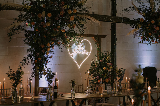 View of wooden arch decorated with wildflowers with couples surname inside love heart light in background